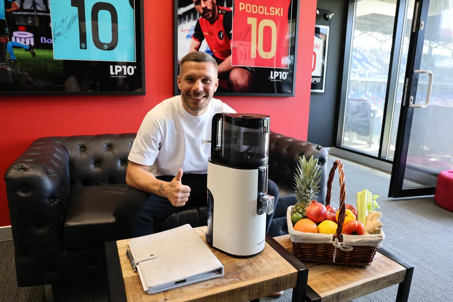 Lukas Podolski z kciukiem w górę na skórzanej kanapie obok białej wyciskarki Hurom i kosza z owocami